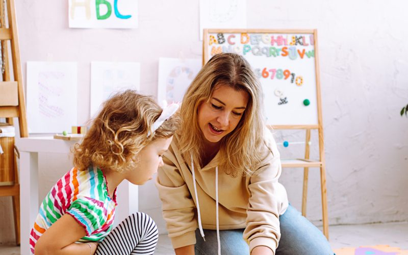 Portrait of curious little girl wear T-shirt, trousers sitting near colorful papers, pencils, different toys with young smiling woman teacher explaining how to draw with finger in classroom. Vertical.
